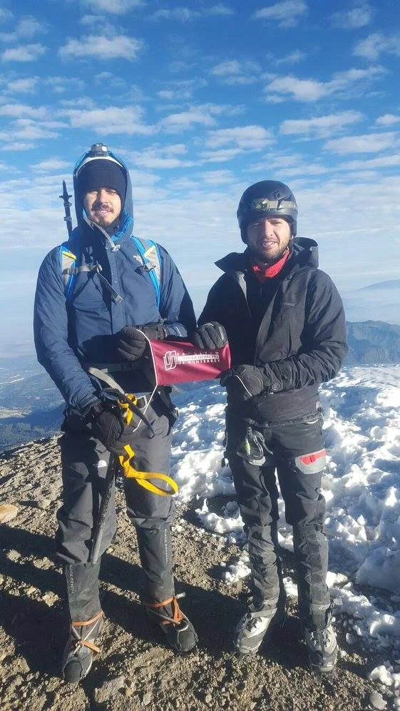 Dos egresados de la FLDM sostienen la bandera de la facultad en la cima de una montaña nevada