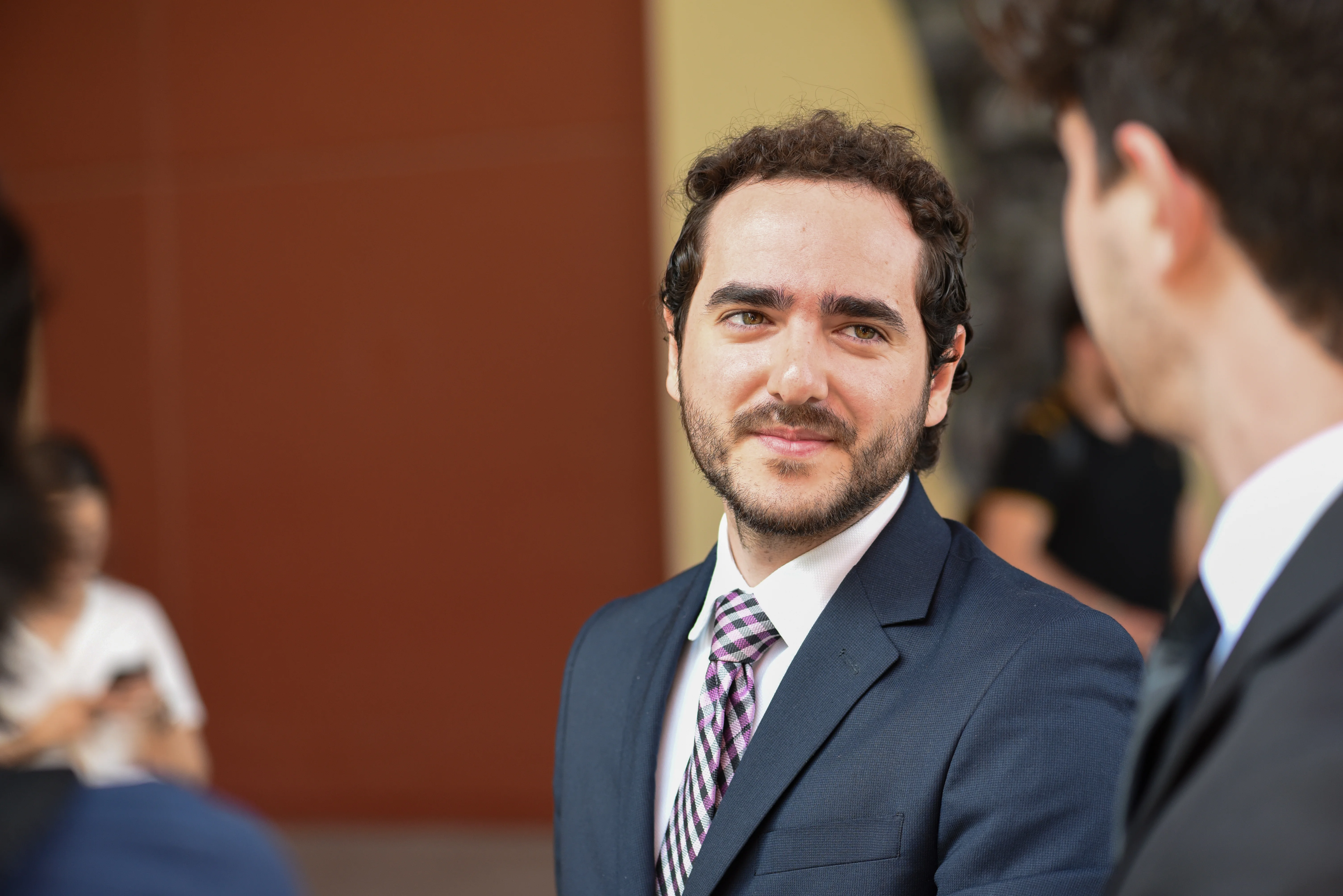 Joven con traje sonrie durante un evento de la FLDM, con companeros de fondo